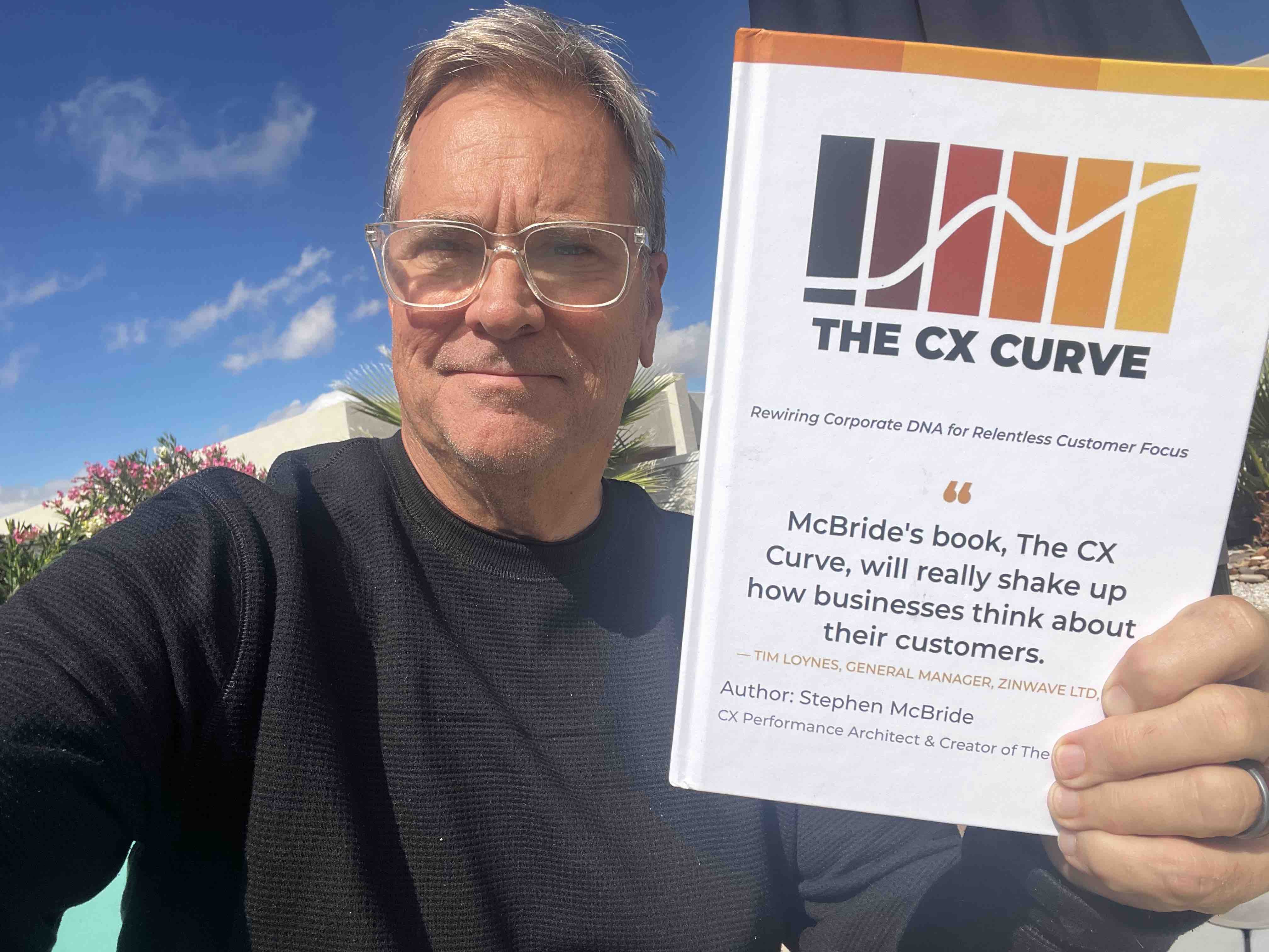 Stephen McBride smiling and holding a copy of his book, The CX Curve, outdoors under a blue sky